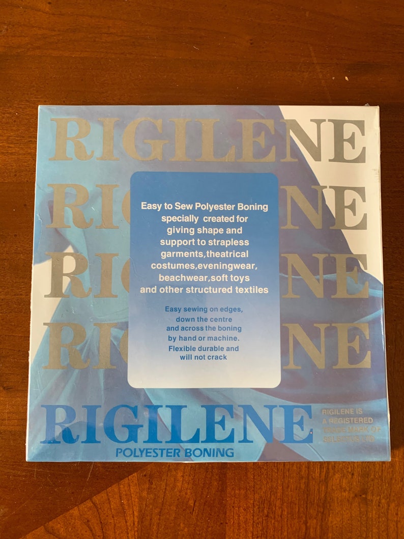K&ouml;nnte beinhalten: Eine blaue und wei&szlig;e Packung Rigilene Polyester-St&uuml;tzstoff. Die Packung enth&auml;lt den Text: "Einfaches N&auml;hen von Polyester-St&uuml;tzstoff, speziell entwickelt f&uuml;r Formgebung und Halt f&uuml;r tr&auml;gerlose Kleidungsst&uuml;cke, Theaterkost&uuml;me, Abendgarderobe, Strandbekleidung, Pl&uuml;schtiere und andere strukturierte Textilien. Einfaches N&auml;hen an den Kanten, unten in der Mitte und &uuml;ber den St&uuml;tzstoff, von Hand oder mit der Maschine. Flexibel, strapazierf&auml;hig und rei&szlig;fest. Rigilene ist eine eingetragene Marke von Selkirk."