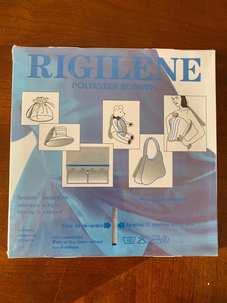 K&ouml;nnte beinhalten: Eine blaue Box mit dem Text "RIGILENE POLYESTER BONING" auf der Vorderseite. Die Box enth&auml;lt 46 Meter (50 Yards) Boning, eine Art versteifendes Material, das in der N&auml;herei und im Handwerk verwendet wird. Die Box enth&auml;lt auch Anweisungen zum Waschen und Trockenreinigen des Bonings.
