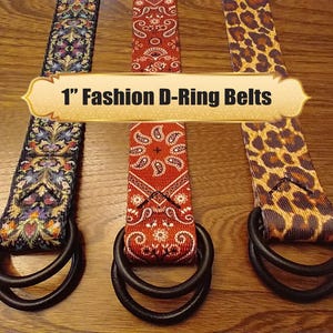May include: Three 1-inch Fashion D-Ring belts with different patterns: floral, paisley, and leopard print. Each belt has a black D-ring buckle. The belts are displayed on a wooden surface.