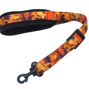 May include: An orange and brown patterned saxophone neck strap with a black padded neck rest and a black hook. The strap features a fall leaf design.