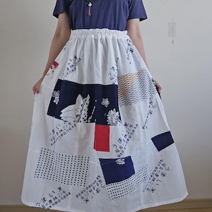 May include: A navy blue t-shirt with a decorative design and a white skirt with patchwork patterns. The skirt features various blue and white designs, including text and geometric shapes. The skirt is a midi length.