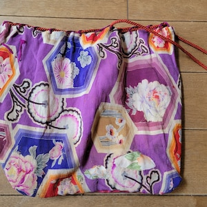 May include: A drawstring bag with a vibrant floral pattern in shades of purple, red, blue, and white. The bag features a repeating hexagonal design with flowers and leaves. A red cord is used for the drawstring.