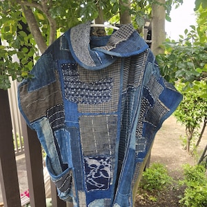 May include: A patchwork jacket in shades of blue, gray, and brown hangs on a white hanger. The jacket features a variety of fabric patterns and textures, including stripes, checks, and solid patches. It has a collar and two pockets. The garment is displayed outdoors.
