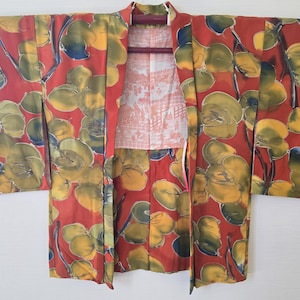 May include: A red kimono with a floral pattern in yellow, green, and blue. The kimono is open, revealing a patterned lining and a wooden hanger. The floral design is large and abstract, covering the entire garment.