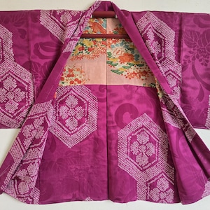 May include: A magenta kimono with a floral pattern on the inside lining. The kimono features geometric designs and a floral pattern. The garment is displayed open, revealing the interior lining with a colorful floral design.