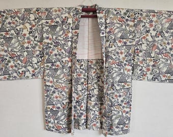 Może przedstawiać: Biały japoński kimono z kwiatowym wzorem w odcieniach niebieskiego, różowego i żółtego. Kimono ma tradycyjny wzór z szerokim kołnierzem i długimi rękawami.