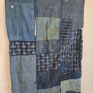 Peut inclure: Textile patchwork aux tons bleu indigo. Le vêtement semble être fait de divers morceaux de tissu, avec des coutures visibles et des zones usées. Le design comprend des motifs géométriques et des pièces bleues unies.