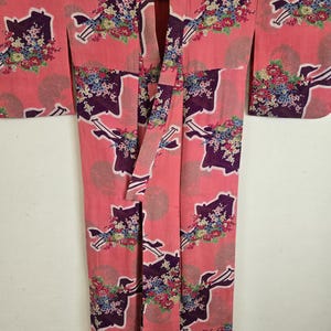 May include: A pink kimono with a repeating pattern of dark purple shapes and floral bouquets. The kimono has long sleeves and a wrap-around design. The floral bouquets feature a mix of colors, including red, blue, and white.