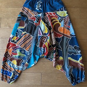 May include: Harem pants featuring a vibrant, multi-colored graphic print with abstract shapes, floral motifs, and Japanese kanji characters. The pants have a solid blue elastic waistband, a low-crotch design, and elasticized cuffs at the ankles.