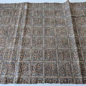 Könnte beinhalten: Ein rechteckiges Stoffstück mit einem sich wiederholenden Gittermuster. Jedes Quadrat enthält ein beiges und braunes Blumenmotiv auf einem gedämpften blaugrauen Hintergrund. Das Design ist von dünnen, dunkelbraunen Linien umrahmt.