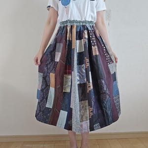 Könnte beinhalten: Ein weißes T-Shirt mit einem blau-weißen Applikationsdesign, kombiniert mit einem Patchwork-Midirock in verschiedenen Farben und Mustern. Das Outfit wird durch rosa-weiß gemusterte Sandalen ergänzt.