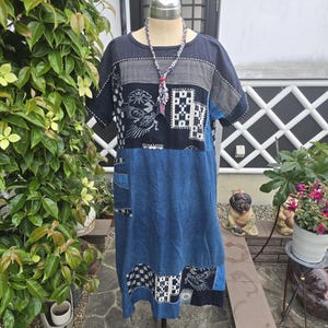 May include: A patchwork dress in shades of blue and navy, displayed on a mannequin. The dress features a round neckline, short sleeves, and a knee-length hem. Decorative patches with geometric and bird designs add visual interest. A beaded necklace completes the look.