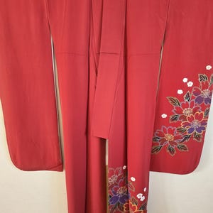 以下が含まれることがあります： 紫、黄色、白の色合いの花柄刺繍が施された赤い着物。着物は滑らかな生地でできており、長い袖が付いています。花柄のデザインは袖と衣服の下部にあります。