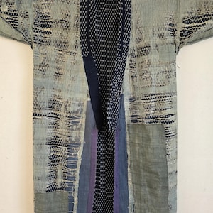 May include: A vintage kimono-style jacket in blue and grey hues. The jacket displays visible mending and patchwork, with a dark blue collar and accents. The fabric has a distressed appearance, with visible wear and tear.