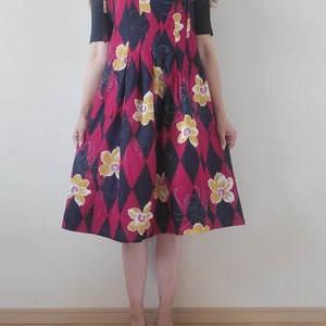 以下が含まれることがあります： 赤と黒のフローラルプリントのサンワンピースに黄色の花が咲いています。ワンピースはスクエアネックラインと細いストラップが付いています。ストラップは肩で結ばれています。