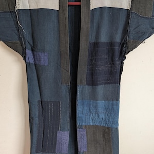 May include: A long-sleeved jacket constructed from various rectangular fabric patches in shades of indigo, navy, charcoal, and light gray. The garment features visible stitching throughout, a dark collar, and a layered, pieced-together design.