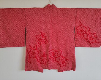 Puede incluir: Un kimono japonés rojo con un patrón floral. El kimono tiene un diseño tradicional con un cuello ancho y mangas largas. El patrón floral es de un tono de rojo más oscuro y está bordado en la tela.