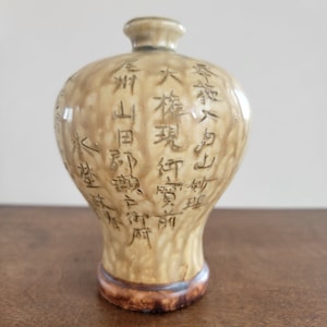 May include: A tan ceramic vase with a rounded body and a narrow neck. The vase is decorated with a pattern of raised, white characters in a traditional Japanese style.