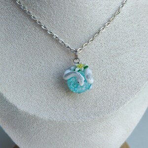 May include: A silver-toned chain necklace featuring a pendant with a small, textured, light blue sphere. The sphere is adorned with two white, curved, animal-like figures and a tiny, pale yellow flower with green leaves attached to the top.