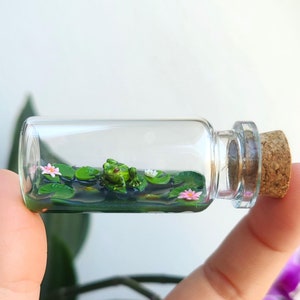 Könnte beinhalten: Eine kleine Glasflasche mit einem Korkverschluss enthält einen winzigen grünen Frosch, der auf einem Bett aus grünen Seerosenblättern mit rosa Blüten sitzt. Die Flasche wird in einer Hand gehalten.
