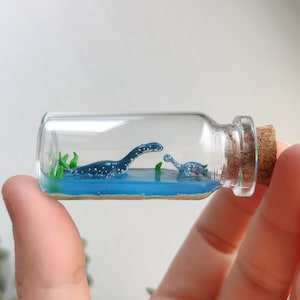 Könnte beinhalten: Eine Glasflasche mit einem Korkverschluss enthält einen winzigen blauen und weißen Dinosaurier und einen kleineren blauen und weißen Dinosaurier in einer blauen Flüssigkeit mit grünen Pflanzen.