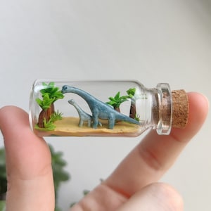Könnte beinhalten: Eine Miniatur-Glasflasche mit einem Korkverschluss enthält einen blauen Dinosaurier und einen kleineren blauen Dinosaurier mit grünen Palmen im Hintergrund.