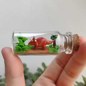 Könnte beinhalten: Eine Miniatur-Figur eines roten Triceratops-Dinosauriers in einer Glasflasche mit einem Korkverschluss. Der Dinosaurier ist von grünen Miniaturpflanzen und einer braunen Basis umgeben.