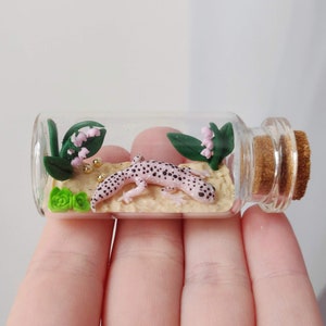 May include: A small glass bottle with a cork stopper contains a pink and black gecko, green leaves, and pink flowers. The bottle is sitting on a hand.