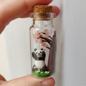 May include: A glass bottle with a cork stopper contains a miniature panda bear sitting under a small pink cherry blossom tree. The panda is made of polymer clay and is painted black and white. The cherry blossom tree is also made of polymer clay and is painted pink and brown.