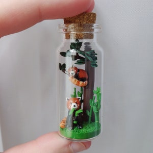 May include: A glass bottle with a cork stopper contains a miniature scene of two red pandas in a forest setting. One red panda is perched on a tree branch, while the other is eating bamboo on the forest floor. The scene is made of polymer clay and is a unique and whimsical piece of art.
