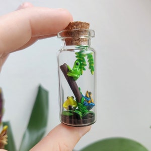 K&ouml;nnte beinhalten: Kleine, transparente Glasflasche mit Korken, gef&uuml;llt mit einem Miniatur-Diorama von drei bunten Fr&ouml;schen. Ein gr&uuml;ner Frosch sitzt auf einem Ast, gelbe und blaue Fr&ouml;sche am Boden, mit gr&uuml;nem Laub.