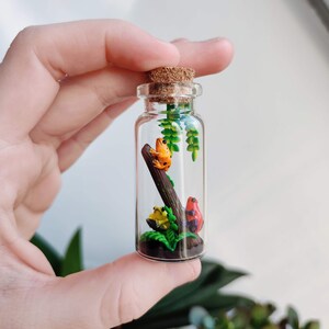 K&ouml;nnte beinhalten: Eine kleine Glasflasche mit Korken, die ein Miniatur-Diorama von bunten Fr&ouml;schen auf einem Ast und Bl&auml;ttern enth&auml;lt. Die Fr&ouml;sche sind orange, gelb und rot mit schwarzen Punkten. Die Flasche ist transparent und zeigt die Szene.