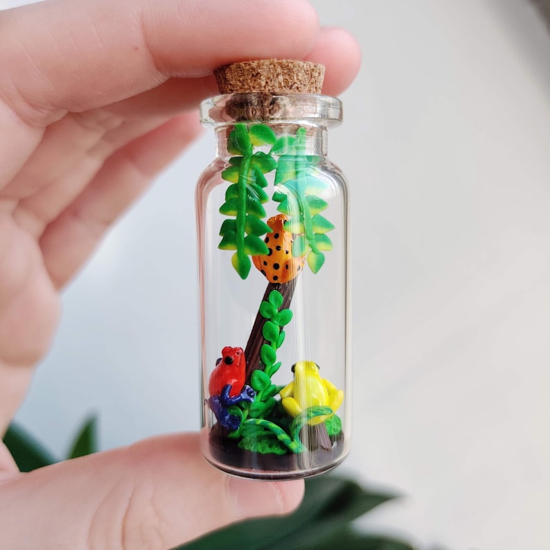 K&ouml;nnte beinhalten: Eine kleine, transparente Glasflasche mit Korken, die ein Miniatur-Diorama enth&auml;lt. Im Inneren befinden sich drei farbenfrohe Froschfiguren, ein Baum und gr&uuml;ne Bl&auml;tter. Die Fr&ouml;sche sind rot, gelb und orange mit schwarzen Punkten.