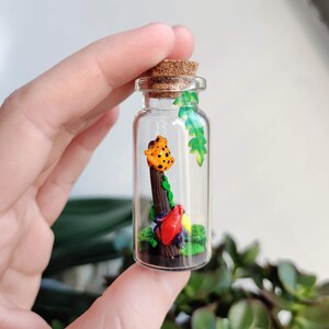 K&ouml;nnte beinhalten: Kleine Glasflasche mit Korken, die eine Miniatur-Szene enth&auml;lt. Ein orangefarbener Frosch mit schwarzen Punkten sitzt auf einem braunen Holzpfosten, umgeben von bunten Bl&auml;ttern. Die Flasche wird in einer Hand gehalten.