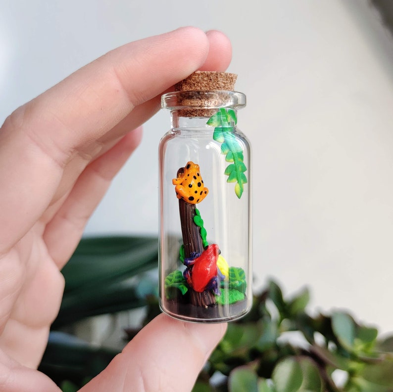 K&ouml;nnte beinhalten: Kleine Glasflasche mit Korken, die eine Miniatur-Szene enth&auml;lt. Ein orangefarbener Frosch mit schwarzen Punkten sitzt auf einem braunen Holzpfosten, umgeben von bunten Bl&auml;ttern. Die Flasche wird in einer Hand gehalten.