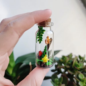 K&ouml;nnte beinhalten: Ein Miniatur-Terrarium in einer durchsichtigen Glasflasche mit Korken. Im Inneren befinden sich farbenfrohe Tonfiguren von Fr&ouml;schen, Pflanzen und Pilzen. Die Flasche wird in einer Hand gehalten, mit einem verschwommenen gr&uuml;nen Hintergrund.
