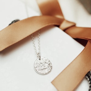 May include: A silver necklace with a round pendant featuring a textured fingerprint design. The necklace is displayed on a white surface with a brown ribbon.