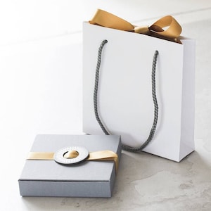 Puede incluir: Una caja de regalo gris con una cinta dorada y un diseño circular en la tapa. La caja está situada junto a una bolsa de regalo blanca con una cinta dorada y asas grises.