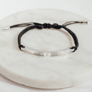 May include: A black braided bracelet with a silver bar engraved with two fingerprints and an ampersand symbol.