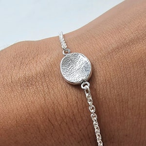 Könnte beinhalten: Ein silbernes Armband mit einem strukturierten, kreisförmigen Anhänger. Der Anhänger ist mit einer filigranen Kette verbunden. Das Armband ist für das Handgelenk konzipiert und wird vor einem Hautton-Hintergrund gezeigt.