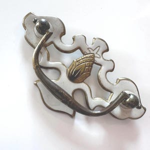 May include: Decorative drawer pull with a white and gold floral design. The pull features a curved handle and a detailed gold-colored accent in the center. The hardware is made of metal and has a vintage aesthetic.