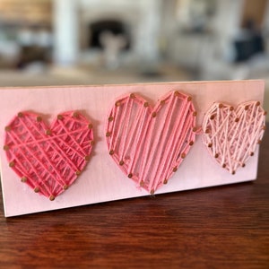 May include: Three pink and white string art hearts on a pink wooden board. The hearts are made with string wrapped around nails.