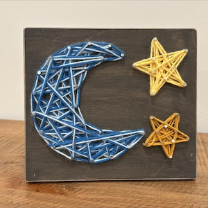 May include: A wooden wall hanging with a blue and white crescent moon and two yellow stars made with string art.
