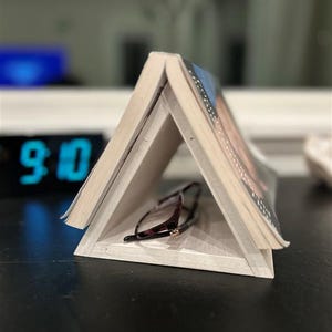 May include: A wooden book stand in a triangular shape, holding a pair of eyeglasses. The stand is made of light-colored wood and the glasses have a dark tortoiseshell pattern. The book is open, with pages visible.