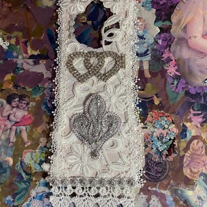 May include: A white lace door hanger with floral and heart embellishments. The door hanger features a silver heart design and a silver fleur-de-lis. The bottom edge has a decorative fringe.