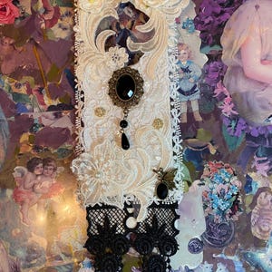 May include: A decorative, rectangular piece featuring cream-colored lace, adorned with a black oval pendant, black lace accents, and small white flowers. A vintage image is centered on the piece.