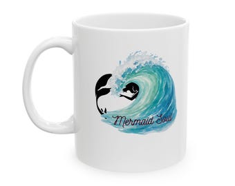Puede incluir: Una taza de cerámica blanca con un gráfico estilo acuarela de una ola oceánica azul y turquesa. Una silueta negra de una sirena es visible dentro de la ola, y el texto Mermaid Soul está escrito en una fuente cursiva oscura debajo.