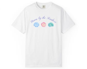 Puede incluir: Una camiseta blanca de manga corta con el texto azul en cursiva Down by the Seashore centrado sobre tres pequeñas ilustraciones estilo acuarela de una vieira azul, un dólar de arena rosa y una concha espiral color verde azulado en el pecho.