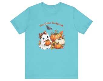 Puede incluir: Una camiseta azul claro con un gráfico de un fantasma blanco con un lazo a cuadros que sostiene una pequeña calabaza. El diseño incluye calabazas naranjas, un girasol, hojas de otoño, un pequeño murciélago y el texto Too Cute To Spook.