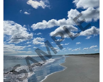 Puede incluir: Una fotografía de una playa de arena bajo un cielo azul brillante con nubes blancas dispersas y un destello solar. Las olas del océano bañan suavemente la orilla, creando una superficie de arena mojada reflectante. La palabra SAMPLE está superpuesta.